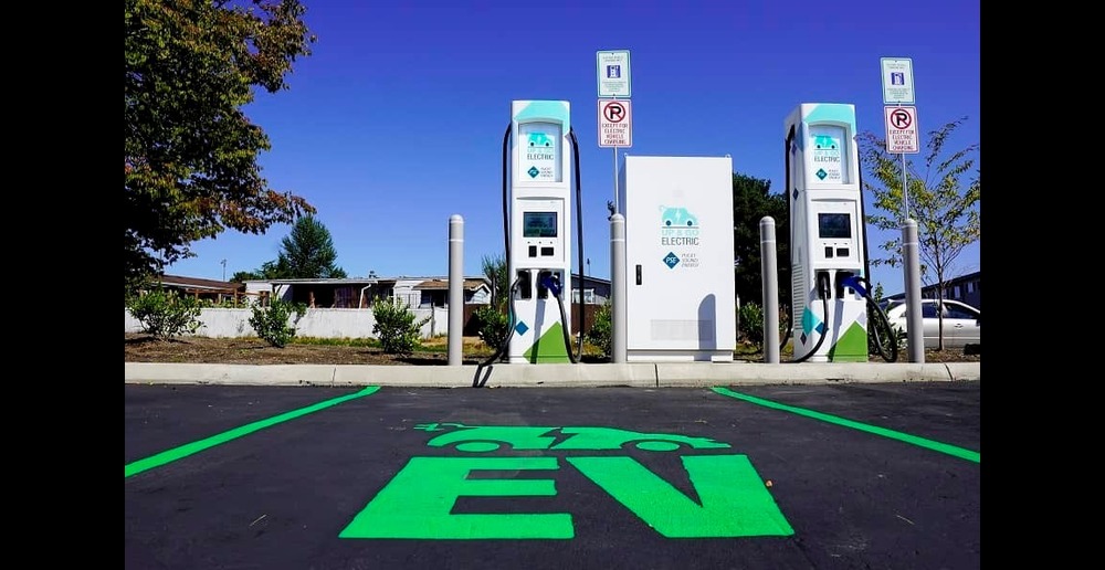 A parking spot with a green electric vehicle logo painted over the black asphalt and two chargers in the backgrounds.